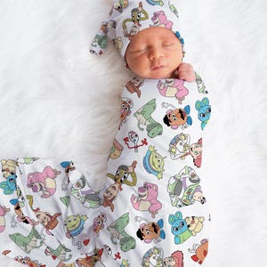 May include: A white swaddle blanket with a pattern of Toy Story characters, including Woody, Buzz Lightyear, Jessie, and Rex. A baby is wearing the swaddle and a matching hat.