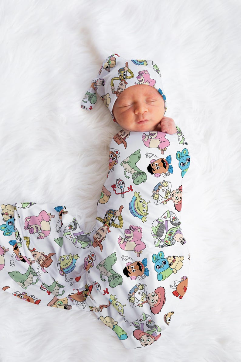 May include: A white swaddle blanket with a pattern of Toy Story characters, including Woody, Buzz Lightyear, Jessie, and Rex. A baby is wearing the swaddle and a matching hat.