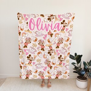 May include: A pink and brown fleece blanket with a repeating pattern of Mickey Mouse and Minnie Mouse characters, teacups, ice cream cones, and rainbows. The blanket has the name "Olivia" written in pink cursive letters.