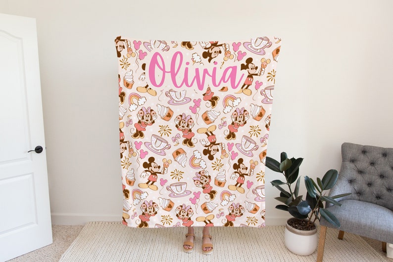 May include: A pink and brown fleece blanket with a repeating pattern of Mickey Mouse and Minnie Mouse characters, teacups, ice cream cones, and rainbows. The blanket has the name "Olivia" written in pink cursive letters.