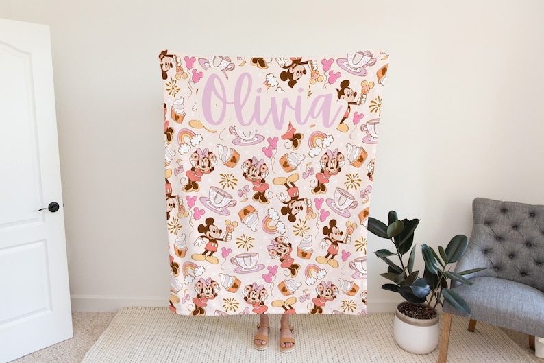 May include: A pink and white fleece blanket with a repeating pattern of Mickey Mouse and Minnie Mouse characters, hearts, cupcakes, and rainbows. The blanket has the name "Olivia" written in pink script.