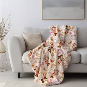 May include: A soft, plush throw blanket featuring a repeating pattern of Mickey Mouse and Minnie Mouse in vintage-inspired outfits. The design includes cupcakes, rainbows, and fireworks on a cream background.