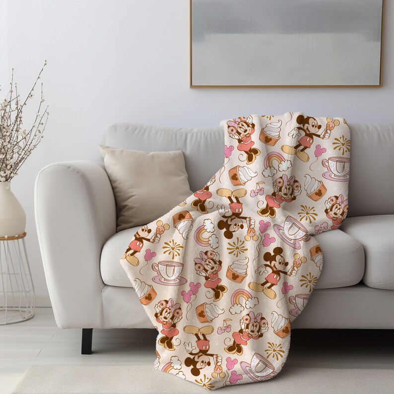 May include: A soft, plush throw blanket featuring a repeating pattern of Mickey Mouse and Minnie Mouse in vintage-inspired outfits. The design includes cupcakes, rainbows, and fireworks on a cream background.