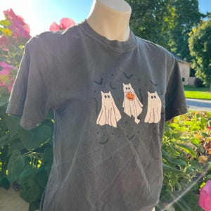 May include: Dark grey t-shirt with a Halloween graphic. The design shows three cartoon ghosts, one with a pumpkin, plus bats and a crescent moon. The shirt has short sleeves and a crew neck.