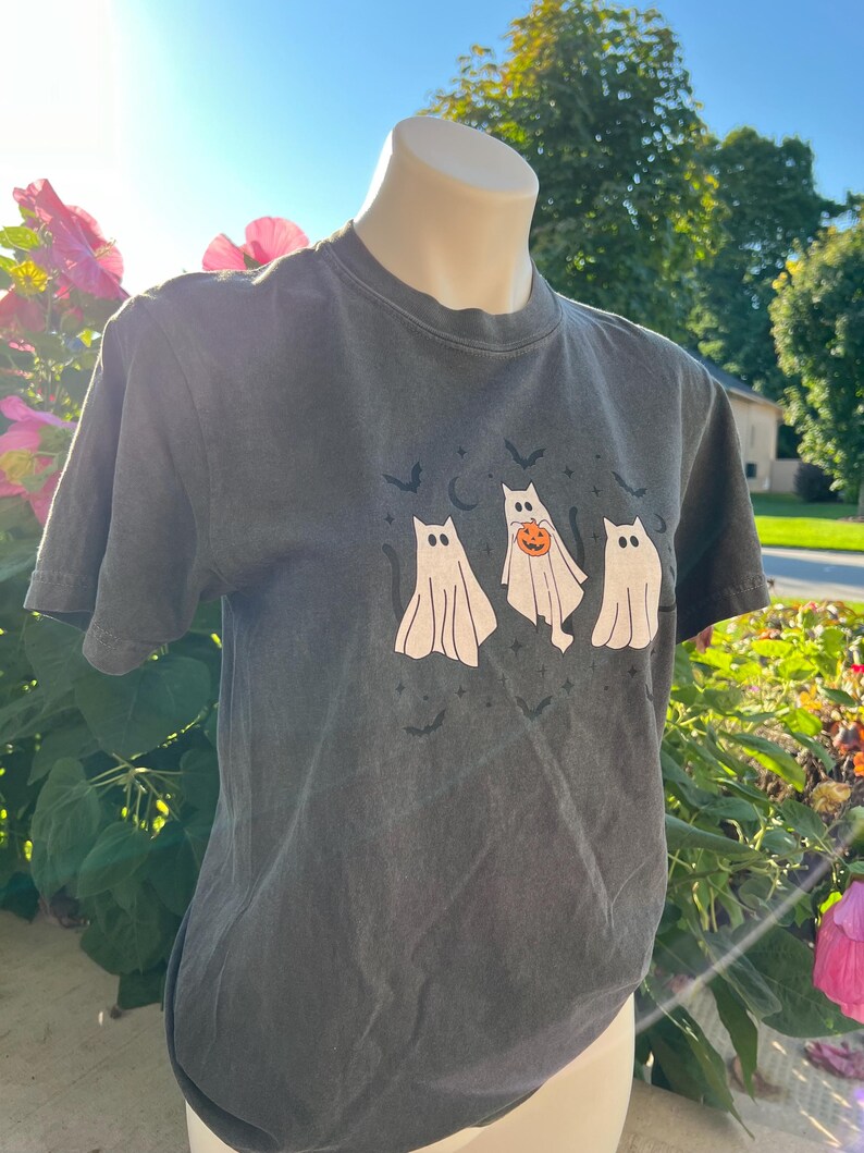 May include: Dark grey t-shirt with a Halloween graphic. The design shows three cartoon ghosts, one with a pumpkin, plus bats and a crescent moon. The shirt has short sleeves and a crew neck.