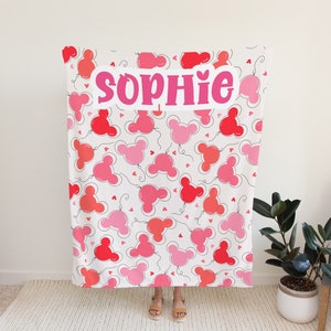 May include: A white fleece blanket with a pink and red balloon pattern. The balloons are shaped like Mickey Mouse ears. The name "Sophie" is written in pink letters across the blanket.