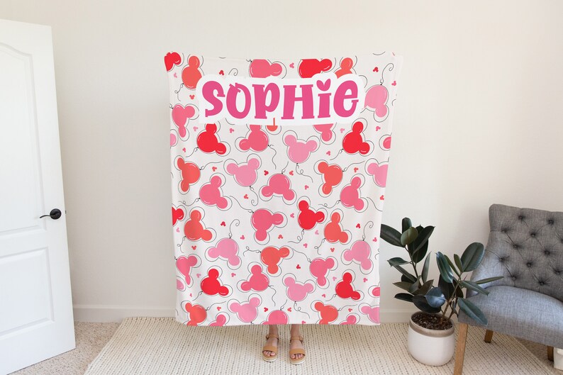May include: A white fleece blanket with a pink and red balloon pattern. The balloons are shaped like Mickey Mouse ears. The name "Sophie" is written in pink letters across the blanket.