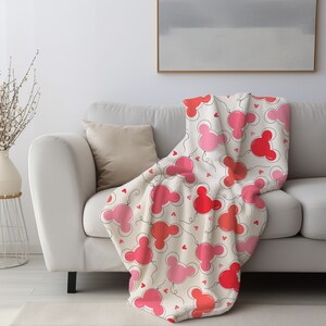 May include: A white fleece throw blanket with a pink and red Mickey Mouse balloon pattern. The blanket is draped over a white couch with a beige throw pillow.