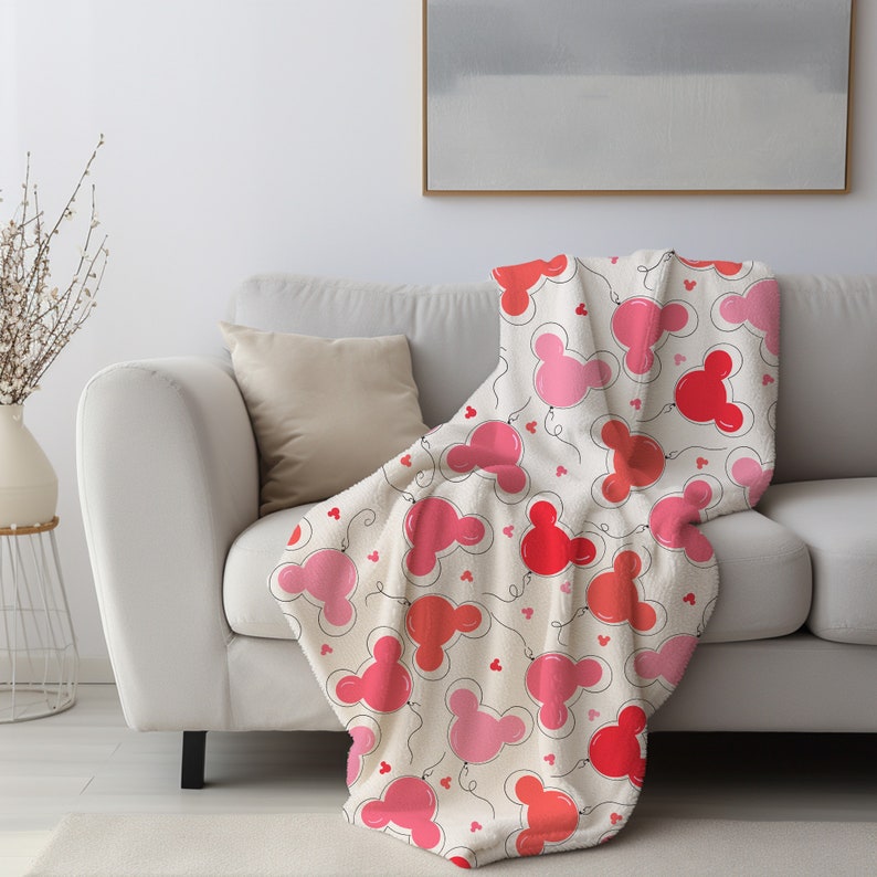 May include: A white fleece throw blanket with a pink and red Mickey Mouse balloon pattern. The blanket is draped over a white couch with a beige throw pillow.