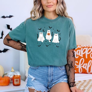 May include: Teal t-shirt with a Halloween ghost design. Three cartoon ghosts are depicted, one with a pumpkin, surrounded by bats and stars. A casual, comfortable style.