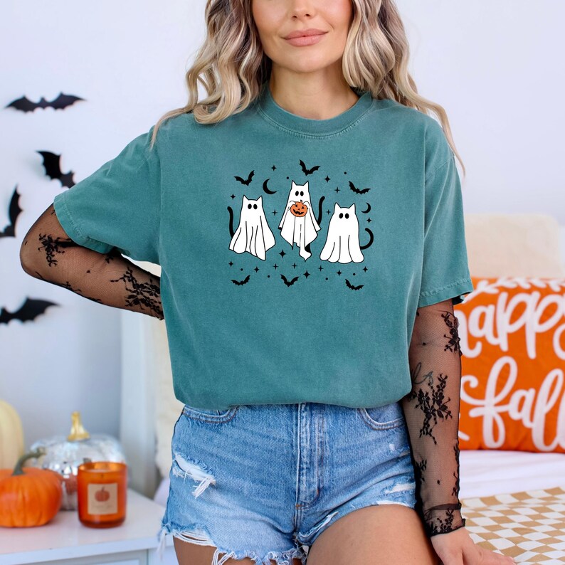 May include: Teal t-shirt with a Halloween ghost design. Three cartoon ghosts are depicted, one with a pumpkin, surrounded by bats and stars. A casual, comfortable style.