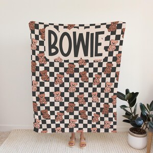 Pu&ograve; includere: Una coperta a scacchi bianco e nero con un motivo di orsacchiotti marroni. La coperta ha la scritta "BOWIE" stampata in lettere nere su sfondo bianco.