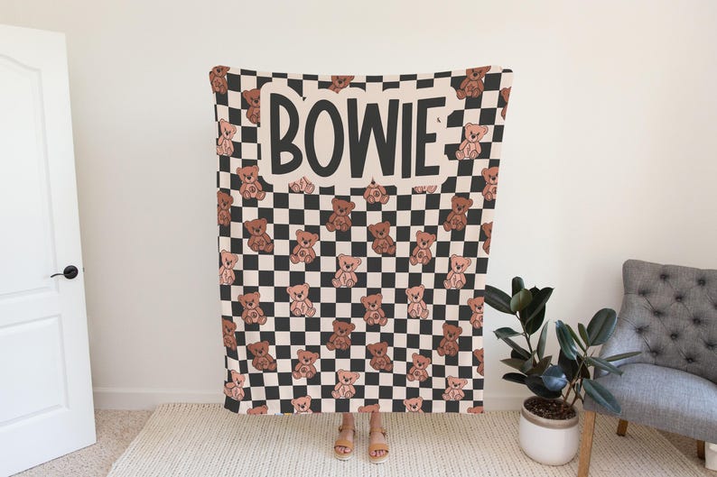 Pu&ograve; includere: Una coperta a scacchi bianco e nero con un motivo di orsacchiotti marroni. La coperta ha la scritta "BOWIE" stampata in lettere nere su sfondo bianco.