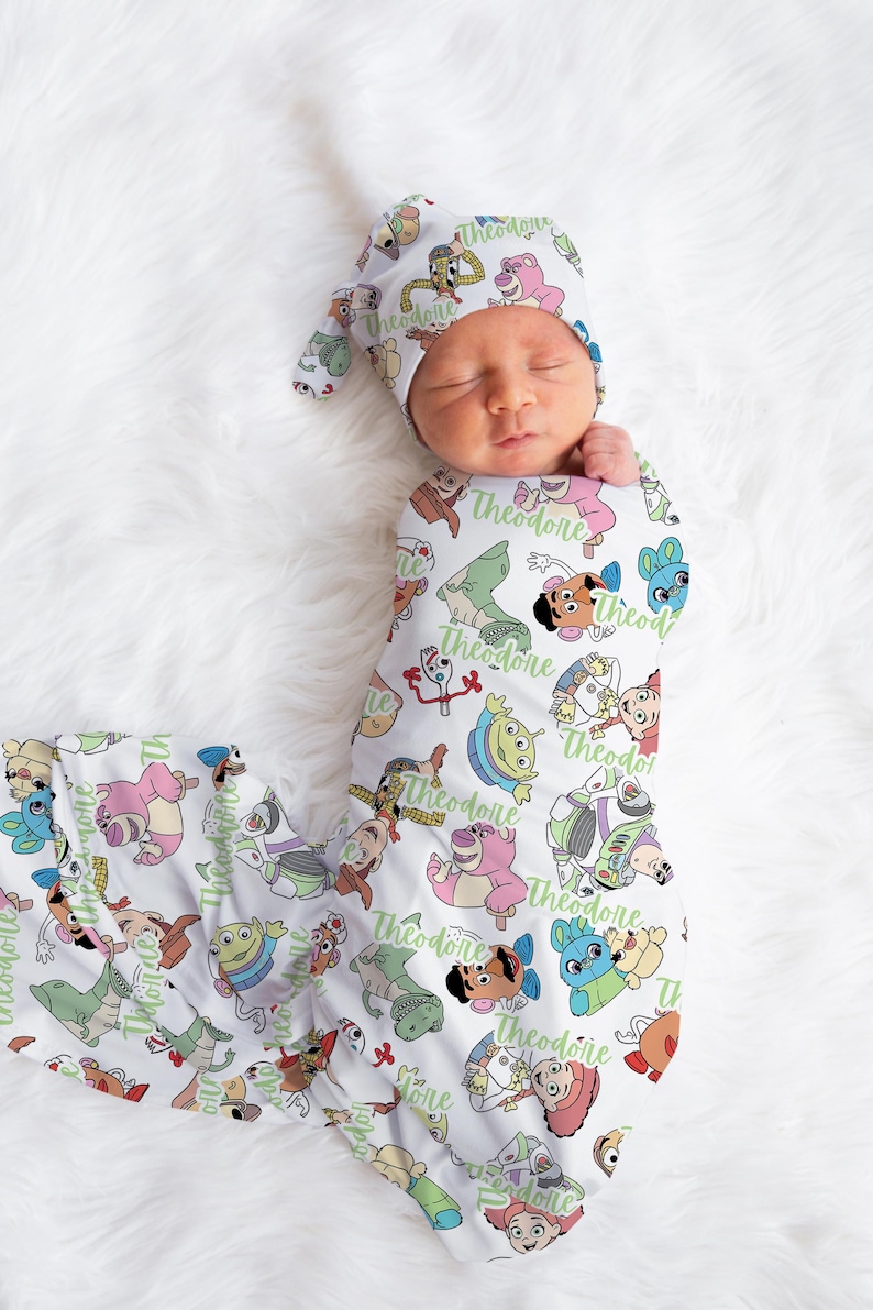 May include: A white swaddle blanket with a repeating pattern of Toy Story characters, including Woody, Buzz Lightyear, and Jessie. The blanket is personalized with the name "Theodore" in green text. A matching hat with the same pattern and personalization is also shown.