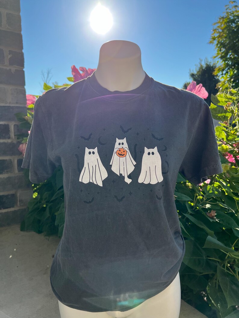 May include: Dark grey t-shirt featuring three cartoon ghosts with cat-like faces. The middle ghost is holding a pumpkin. Small bats are printed above the ghosts. The shirt is displayed on a mannequin in front of a sunny, blue sky.