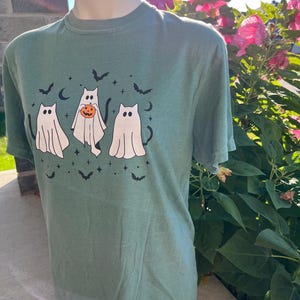 May include: Green t-shirt with a Halloween design. The graphic shows three ghost cats, bats, stars, and crescent moons. One cat holds a pumpkin. The shirt is displayed on a mannequin in front of a brick wall and pink flowers.