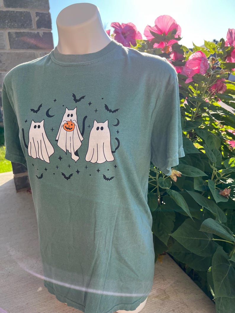 May include: Green t-shirt with a Halloween design. The graphic shows three ghost cats, bats, stars, and crescent moons. One cat holds a pumpkin. The shirt is displayed on a mannequin in front of a brick wall and pink flowers.
