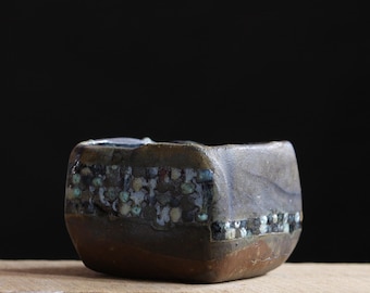 May include: A small, square ceramic bowl with a unique, textured design. The bowl features a dark brown base, a band of speckled blue and white, and a gray top. It sits on a wooden surface against a black background.