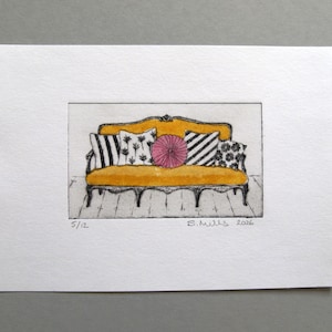 May include: A small ink and watercolour drawing on white paper depicting a yellow sofa with black and white patterned throw pillows and a central round pink cushion. The artwork is signed S. Mills 2026 and numbered 5/12 at the bottom.