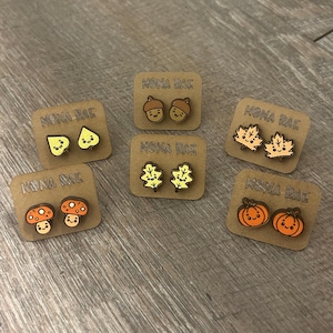 May include: A collection of wooden stud earrings featuring fall-themed designs. The earrings include acorns, leaves, mushrooms, and pumpkins, each with a smiling face. The earrings are displayed on small brown cards with the text "MONA RAE".