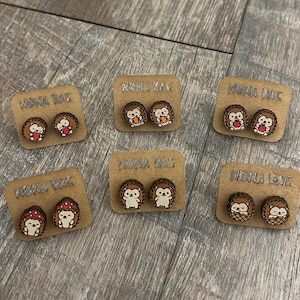 May include: Wooden stud earrings featuring hedgehog designs. Each pair is mounted on a small, rectangular cardboard display with the text "NONA RAE". The earrings showcase various hedgehog illustrations, including those with hearts, baskets, and mushrooms.