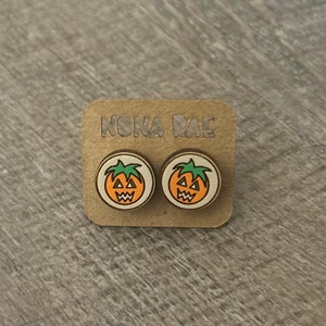 Op de afbeelding: Een paar houten pompoen oorbellen met een jack-o'-lantern gezicht op elke oorbel. De oorbellen zijn op een kaart die "NONA RAE" zegt.