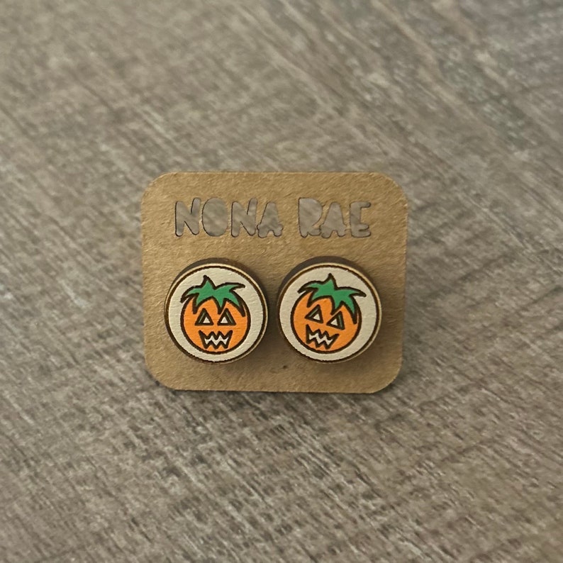Op de afbeelding: Een paar houten pompoen oorbellen met een jack-o'-lantern gezicht op elke oorbel. De oorbellen zijn op een kaart die "NONA RAE" zegt.