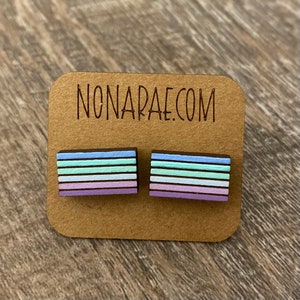 May include: A pair of wooden earrings with a striped design in shades of purple, blue, and green. The earrings are displayed on a brown card with the text "NONARAE.COM".