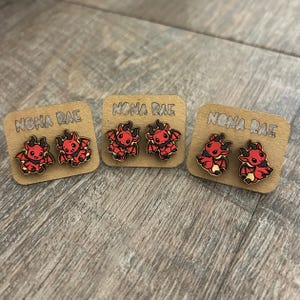 May include: Three pairs of wooden devil-themed earrings on display. Each pair is mounted on a small, rectangular cardboard backing with the text "NONA RAE" printed on it. The earrings feature a red devil design with black accents.