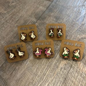 May include: Five pairs of wooden earrings featuring cartoon geese in various outfits. Each pair is mounted on a small, brown card with the text "Nona Rae". The geese are depicted in different poses and wearing hats, sunglasses, and swim trunks.