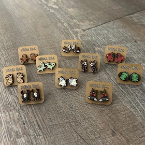 May include: A collection of 10 pairs of wooden earrings featuring various cartoon characters. The earrings are displayed on small cards with the text "NOMA RAE" printed on them.