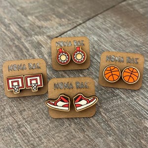 May include: Four pairs of wooden earrings with a basketball theme. The earrings feature a basketball hoop, a basketball, a basketball shoe, and a basketball with a net. Each pair of earrings is displayed on a small wooden card with the text "NONA RAE" printed on it.
