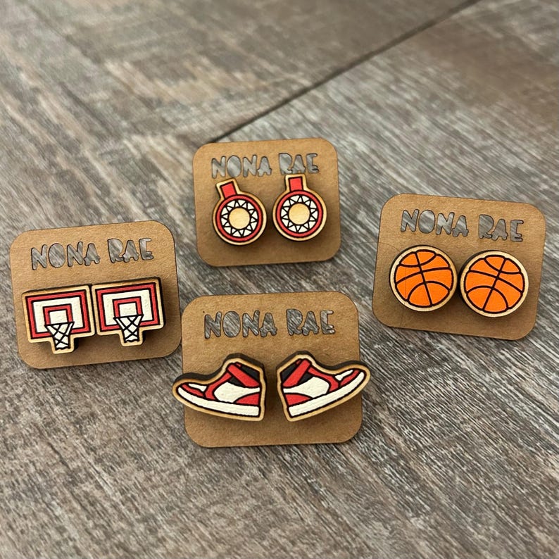May include: Four pairs of wooden earrings with a basketball theme. The earrings feature a basketball hoop, a basketball, a basketball shoe, and a basketball with a net. Each pair of earrings is displayed on a small wooden card with the text "NONA RAE" printed on it.