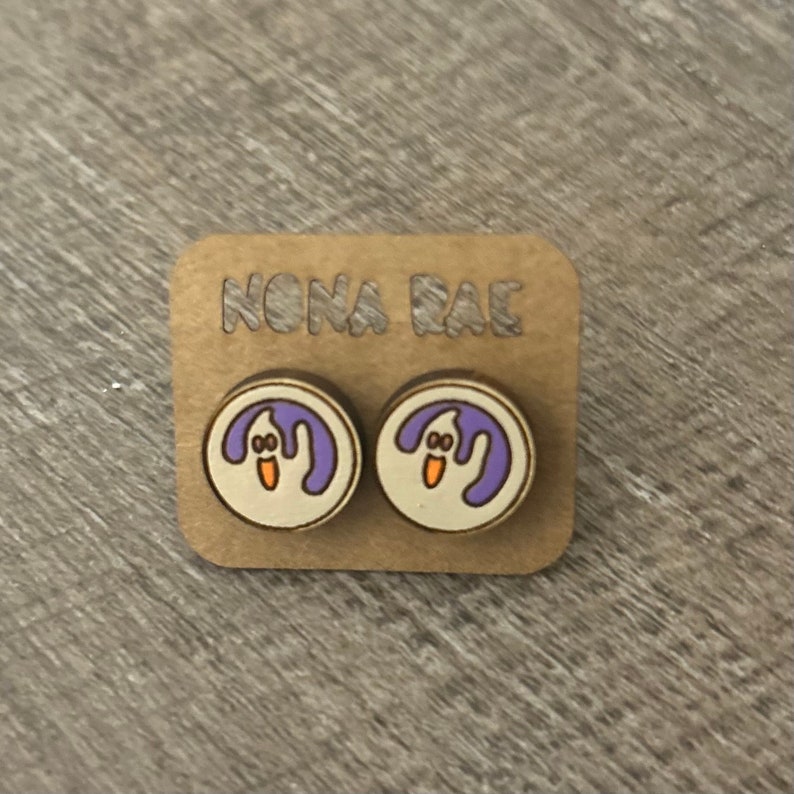 Op de afbeelding: Een paar houten oorbelstekers met een paars spookontwerp. De oorbellen bevinden zich op een kaart met de tekst "NONA RAE".