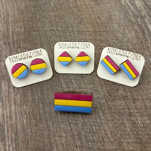 May include: Three sets of earrings and a pin featuring the pansexual pride flag colors: pink, yellow, and blue. The earrings are in the shape of a circle and a diamond. The pin is rectangular.