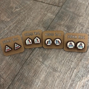 May include: Four pairs of wooden stud earrings on brown cards. Each pair features a different design: a triangle with a book, a house with a book, books inside a circle, and a stack of books with a plant. The cards are labeled "NONA RAE".