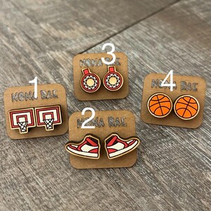 May include: Four pairs of wooden earrings with a basketball theme. The earrings feature basketballs, basketball shoes, basketball hoops, and basketballs with red and white accents. The earrings are labeled with numbers 1 through 4.