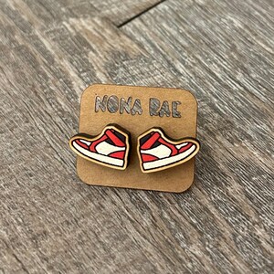 May include: A pair of wooden earrings shaped like red and white sneakers. The earrings are on a brown card with the text "NONA RAE" printed on it.