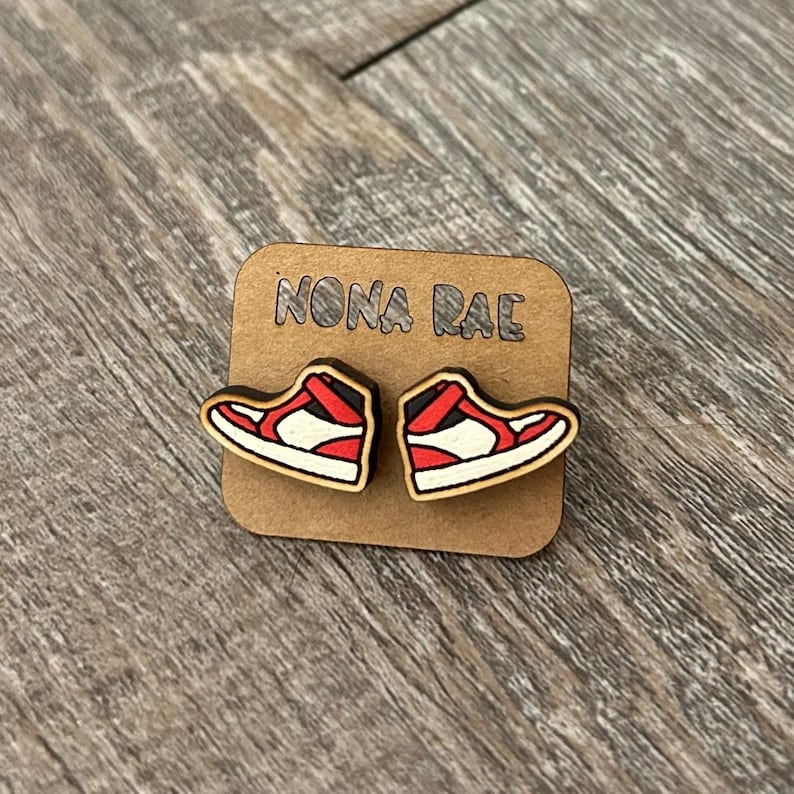 May include: A pair of wooden earrings shaped like red and white sneakers. The earrings are on a brown card with the text "NONA RAE" printed on it.