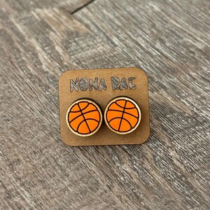 May include: A pair of wooden earrings with a basketball design. The earrings are orange and black and have the text "NONA RAE" on the card they are displayed on.
