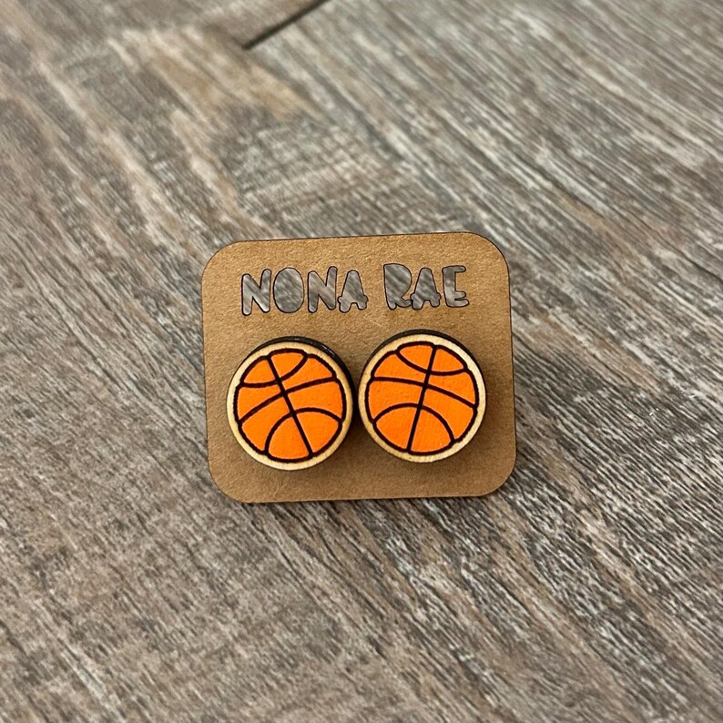 May include: A pair of wooden earrings with a basketball design. The earrings are orange and black and have the text "NONA RAE" on the card they are displayed on.
