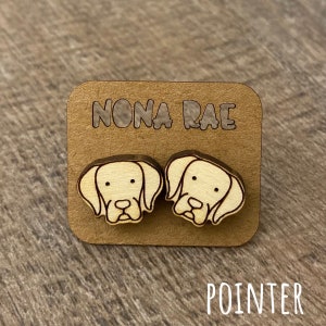 May include: A pair of wooden earrings featuring a light brown pointer dog design. The earrings are on a brown card with the text "NONA RAE" and "POINTER" printed on it.