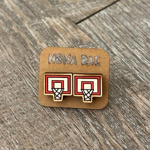 May include: A pair of wooden earrings featuring a basketball hoop design. The hoops are red and white with a black net. The earrings are displayed on a brown card with the text "NONA RAE" printed on it.