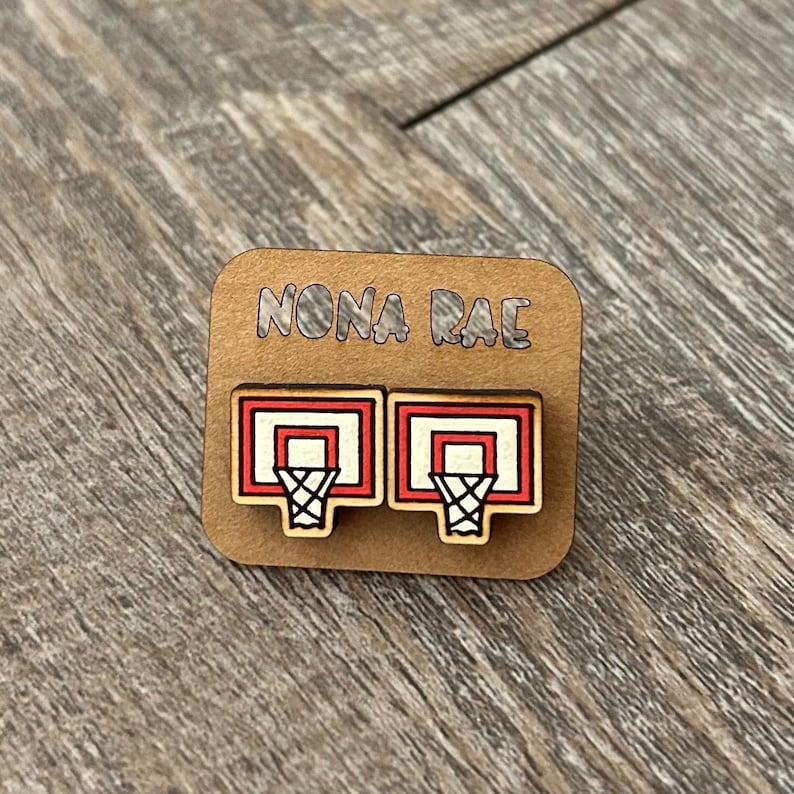 May include: A pair of wooden earrings featuring a basketball hoop design. The hoops are red and white with a black net. The earrings are displayed on a brown card with the text "NONA RAE" printed on it.