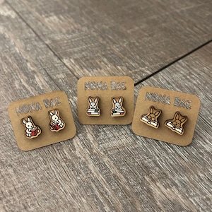 May include: Wooden stud earrings featuring bunny designs. Each pair of earrings is mounted on a small, rectangular cardboard backing with the text "NONA RAE". The bunny designs vary, with some bunnies reading books and others with different poses.