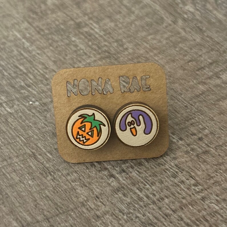 Op de afbeelding: Een paar houten Halloween-oorbellen met een pompoen- en een spookontwerp. De oorbellen bevinden zich op een kaart met de tekst "NONA RAE".