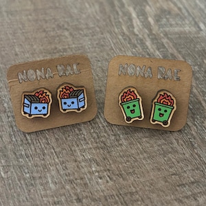 May include: Two pairs of wooden earrings featuring a cartoon dumpster with flames on top. One pair is blue and the other is green. The earrings are displayed on a wooden card with the text "NONA RAE" printed on it.