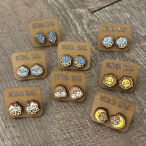 May include: Wooden stud earrings with weather-themed designs, including clouds, rainbows, suns, and moons. Each pair is mounted on a small brown card with the text "NONA RAE". The earrings are painted in various colors, such as blue, yellow, and orange.