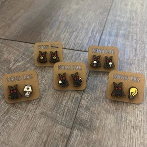 May include: Five pairs of wooden stud earrings on brown cards. Each card has the text "NONA RAE". The earrings feature a black moth design with red eyes, paired with a lamp, lightbulb, or other objects. The earrings are on a wooden surface.