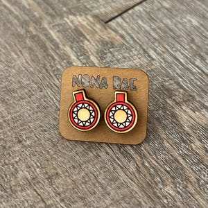 May include: A pair of wooden earrings with a red and white sun design. The earrings are on a card that says "Nona Rae".